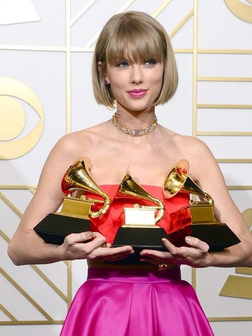 Taylor Swift steht im schulterfreien purpurfarbenen Kleid vor einer weißen Stellwand und hält die drei Grammys in den Händen, die jeweils ein Grammophon darstellen.