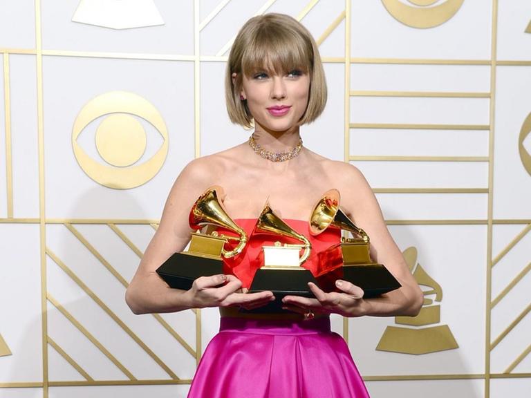 Taylor Swift steht im schulterfreien purpurfarbenen Kleid vor einer weißen Stellwand und hält die drei Grammys in den Händen, die jeweils ein Grammophon darstellen.