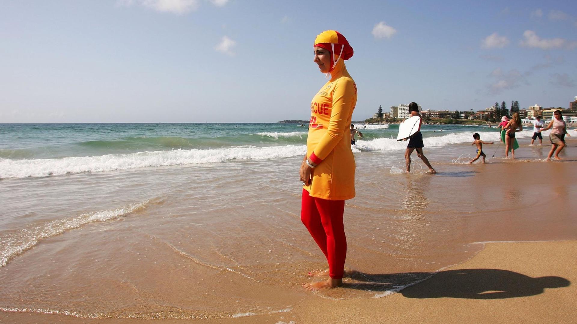 Eine muslimische Rettungsschwimmerin steht in einem Burkini am Strand von Sydney und schaut aufs Meer. Der Burkini ist rot und gelb. Im Hintergrund gehen Menschen mit Surfbrettern ins Wasser.