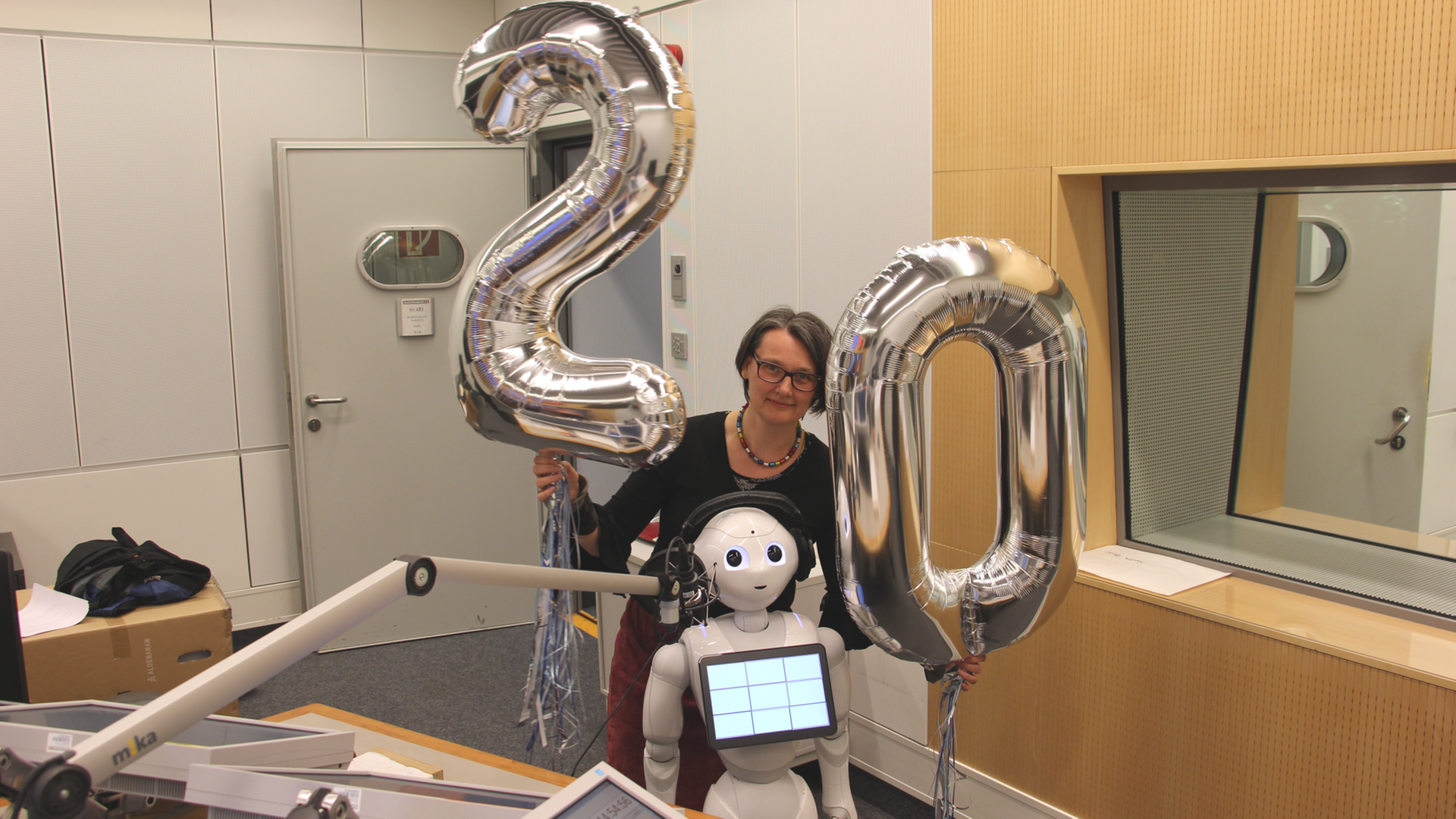 Eine Frau und ein kleiner weißer Roboter stehen im Sendestudio, die Frau hält zwei Ballons in Form einer "20" in den Händen.
