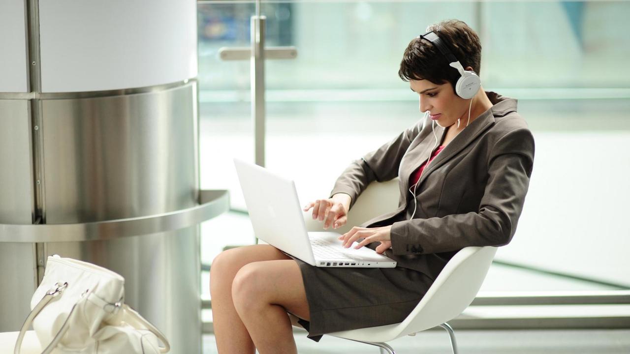 Junge Geschäftsfrau mit sitzt gekrümmt mit einem Laptop auf dem Schoß in einem Büro auf einem Stuhl.