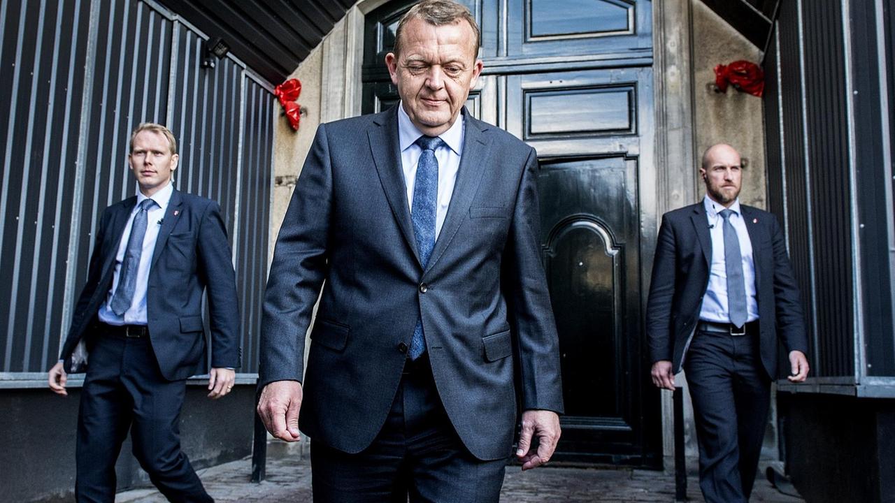 Rasmussen entfernt sich im schwarzen Anzug von einer Tür, rechts und links neben ihm laufen zwei Personenschützer.
