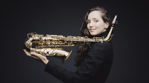 Eine junge Frau, mit langen braunen Haaren, steht seitlich vor einen schwarzen Hintergrund. In der Hand hält sie ihr Saxofon, der obere Teil des Instruments ist auf ihrer Schulter abgelegt.