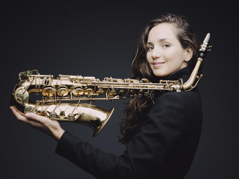 Eine junge Frau, mit langen braunen Haaren, steht seitlich vor einen schwarzen Hintergrund. In der Hand hält sie ihr Saxofon, der obere Teil des Instruments ist auf ihrer Schulter abgelegt.