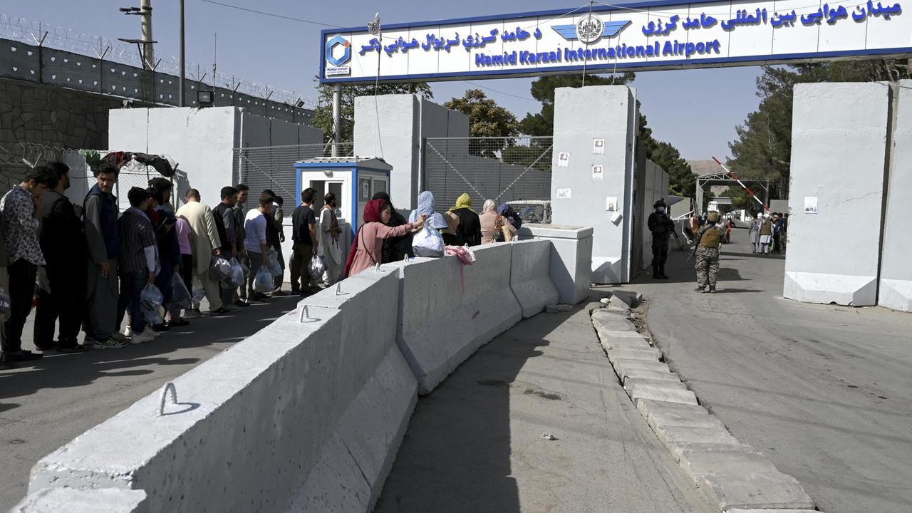 Menschen stehen in einer Schlange vor dem Haupteingang des Kabuler Flughafens. Auf einem Schild steht "Hamid Karzai International Airport".