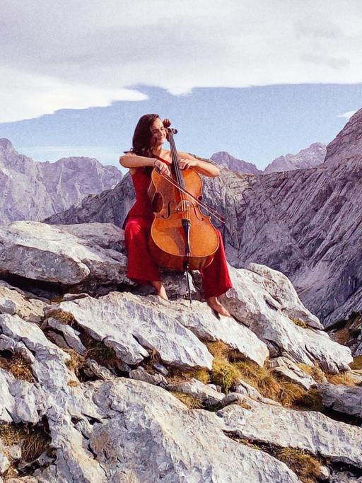 Raphaela Gromes sitzt mit ihrem Cello auf einem Felsen. Im Hintergrund ist ein Bergpanorama zu sehen