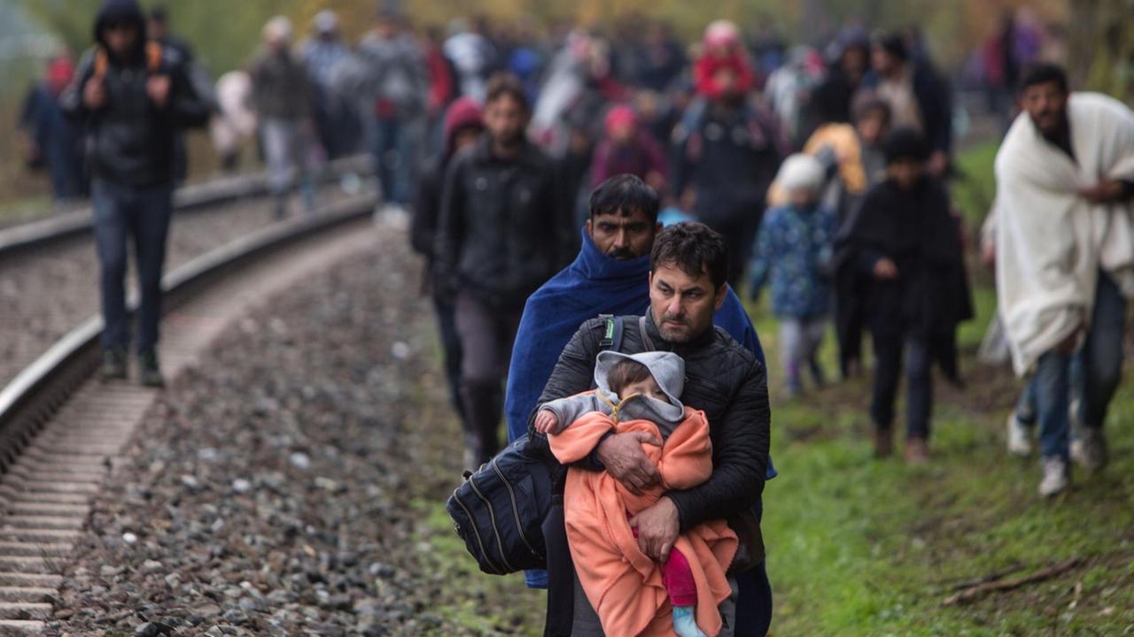 Viele Flüchtlinge laufen auf und entlang einer Bahnstrecke. Im Vordergrund trägt ein Mann ein in eine rote Decke eingewickeltes Kleinkind.