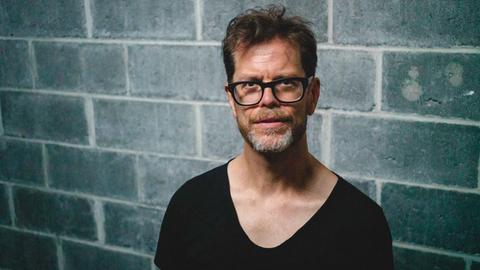 Vor einer Mauer steht ein bebrillter Mann mit tief ausgeschnittenem, schwarzem T-Shirt und schaut in die Kamera.