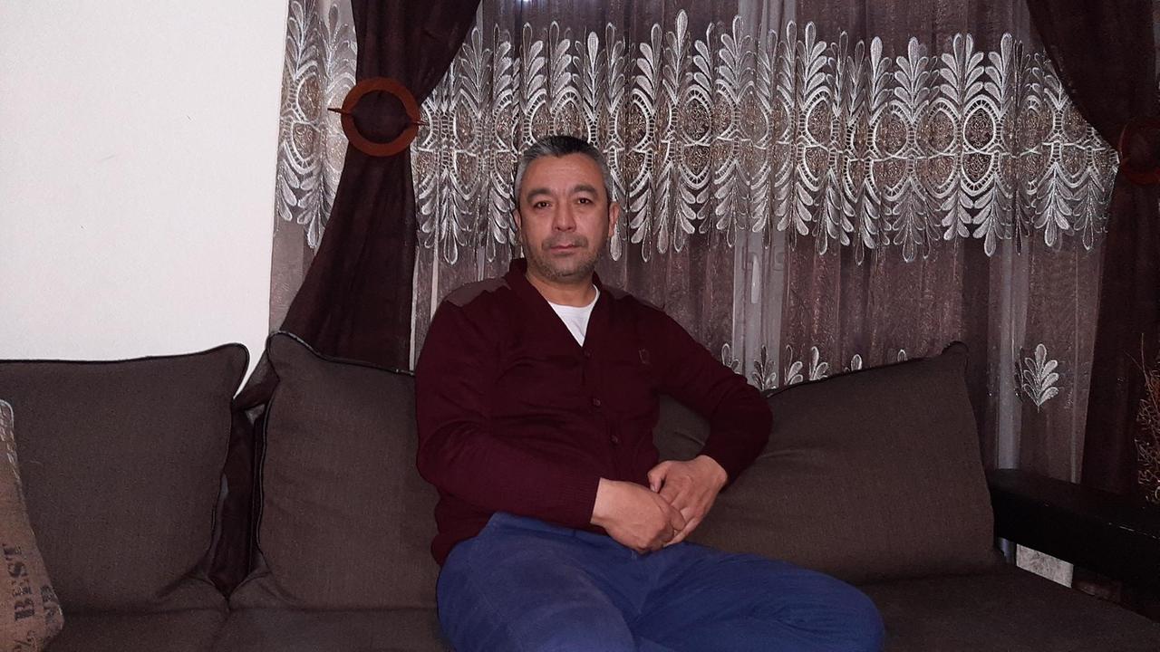 Ein Mann sitzt mit verschränkten Händen in Jeans und Pulli auf einem dunkelbraunen Sofa in einer Wohnung.