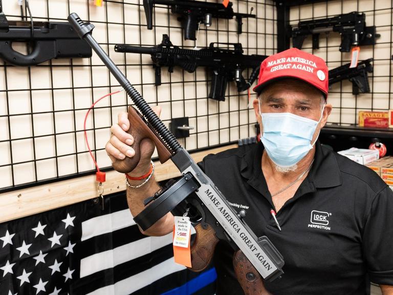 Ein Mitarbeiter eines Waffengeschäfts, mit Gesichtsmaske, präsentiert ein Gewehr. Auf der Waffe und auf der roten Basecap des Verkäufers ist der Schriftzug "Make America Great Again" zu sehen.