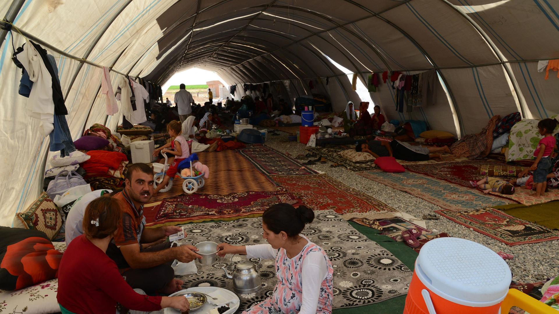 Eine jesidische Familie hat Schutz gefunden in einem Flüchtlingscamp in der Nähe der türkisch-irakischen Grenze. Erwachsene und Kinder sitzen auf ausgebreiteten Teppichen in einem Zelttunnel.