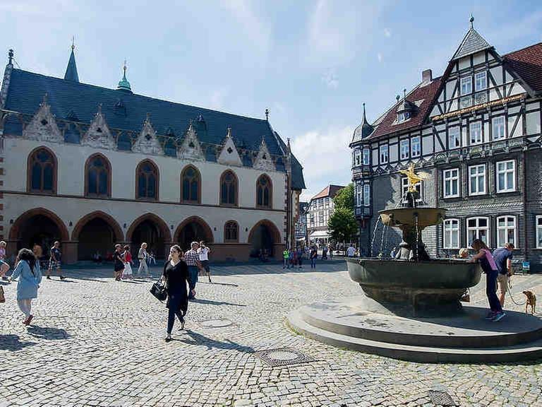 Passanten gehen in Goslar (Niedersachsen) auf dem Marktplatz am Marktbrunnen vorbei. Links ist das Goslarer Rathaus zu sehen.