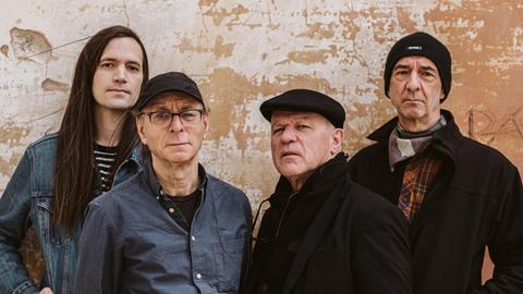 Vier Männer stehen vor einer okerfarbenen Wand. Zusammen sind sie die Band Wire.
