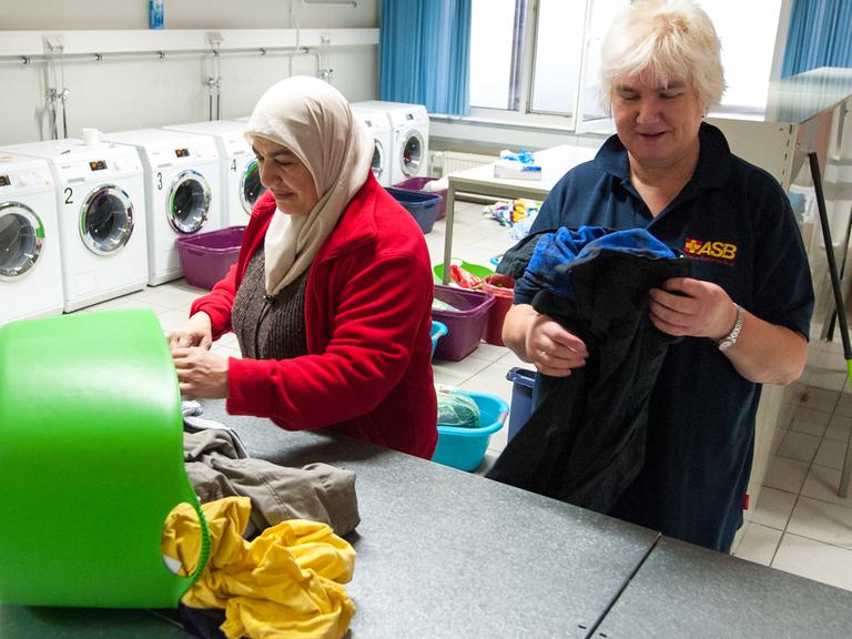 Eine Dame legt zusammen mit einer Flüchtlingsfrau Kleidung in einer Wäscherei zusammen.