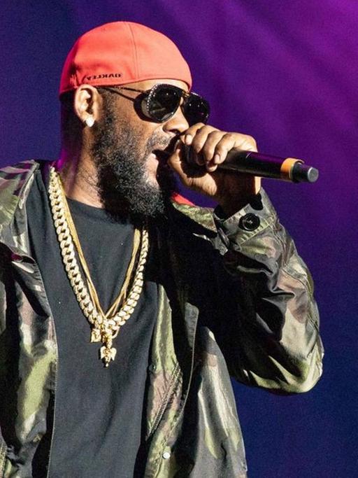 R. Kelly rappt in ein Mikrofon, in seiner Hand trägt er ein Whiskeyglas und Zigarre.