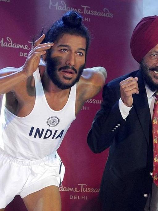 Milkha Singh steht freudig lachend neben seiner Wachsfigur, die ihn als jungen Laufsportler zeigt.
