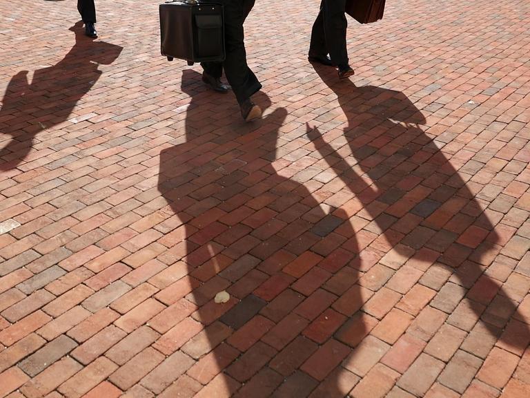 Personen mit Koffern werfen Schatten auf ein rotes Straßenpflaster.