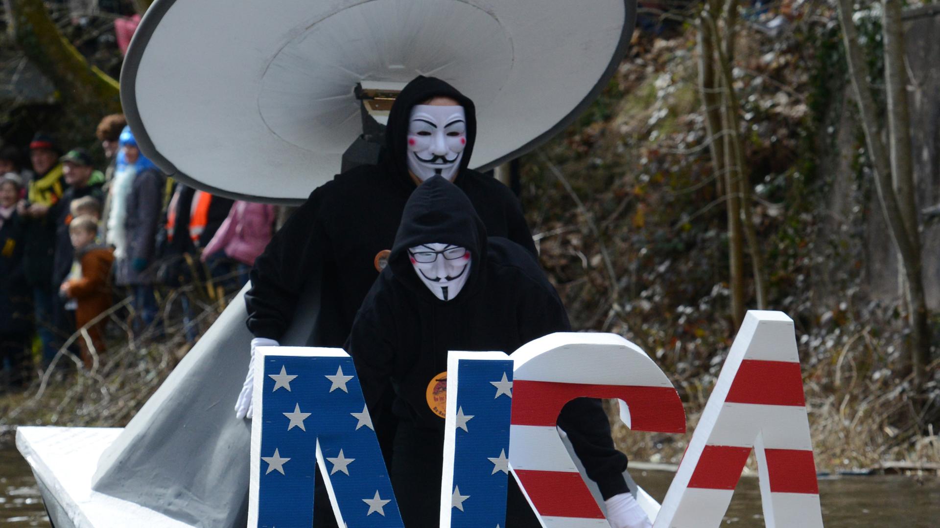 Zwei Männer auf einem Boot, verkleidet als Guy Fawkes mit weißer Maske, fahren einen Bach hinunter, vor ihnen stehen die fast mannshohen Buchstaben NSA in den Farben der amerikanischen Flagge