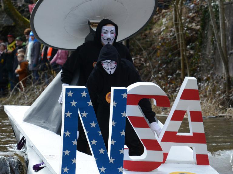 Zwei Männer auf einem Boot, verkleidet als Guy Fawkes mit weißer Maske, fahren einen Bach hinunter, vor ihnen stehen die fast mannshohen Buchstaben NSA in den Farben der amerikanischen Flagge