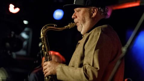 Profilansicht eines etwa 60-jährigen Mannes mit Baseballkappe und beigem Jackett, der Tenorsaxofon spielt