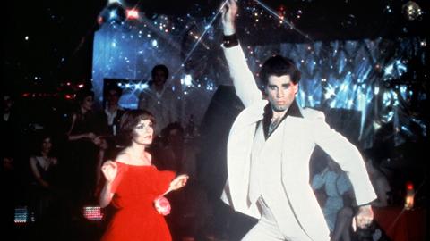 Karen Lynn Gorney und John Travolta in einer Szene aus "Saturday Night Fever", beide tanzen in einer Disco, John Travolta reckt den rechten Arm in die Höhe und biegt die Hüfte durch.