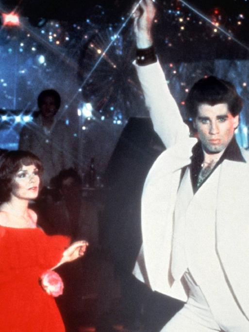Karen Lynn Gorney und John Travolta in einer Szene aus "Saturday Night Fever", beide tanzen in einer Disco, John Travolta reckt den rechten Arm in die Höhe und biegt die Hüfte durch.