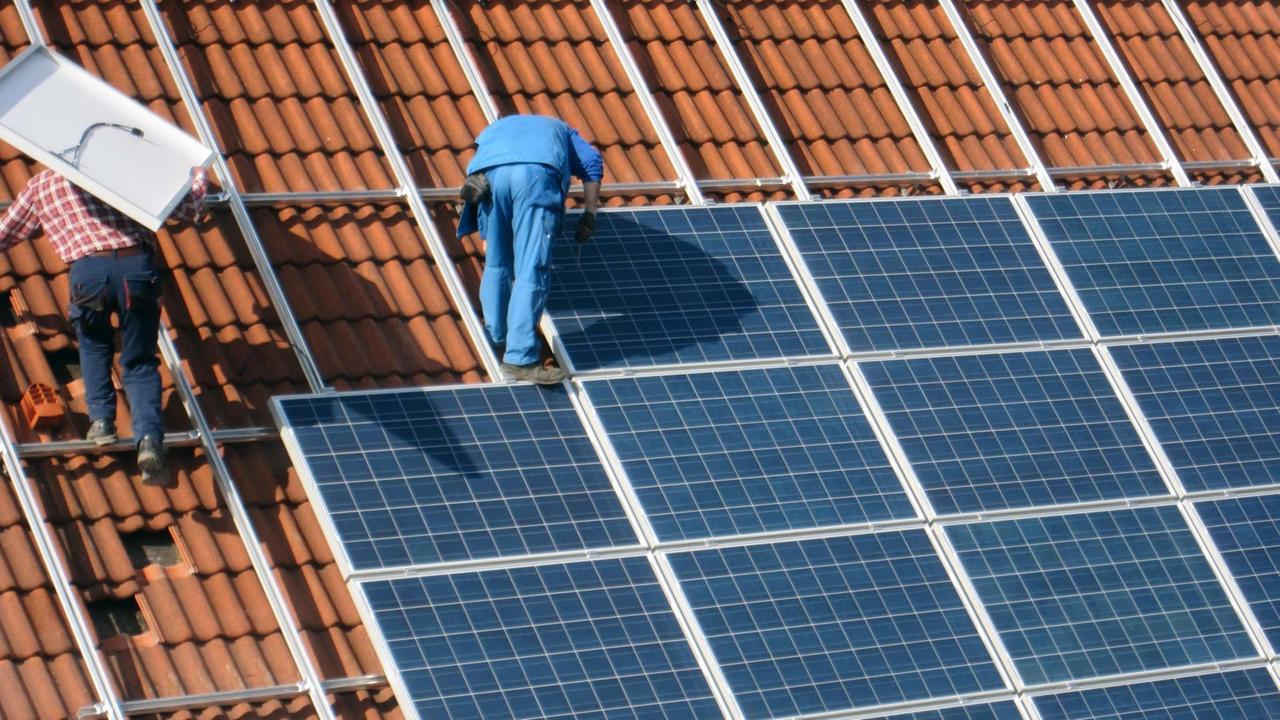 Zwei Arbeiter montieren Solarzellen auf einem roten Ziegeldach.