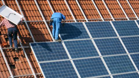 Zwei Arbeiter montieren Solarzellen auf einem roten Ziegeldach.