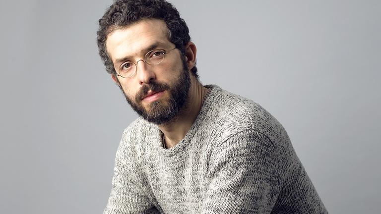 Omer Meir Wellber sitzt auf einem Hocker, trägt einen grauen Strickpullover und blickt in die Kamera