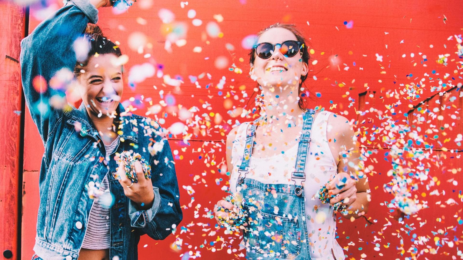 Zwei Frauen stehen vor einer roten Wand und werfen voll Freude Konfetti in die Luft.