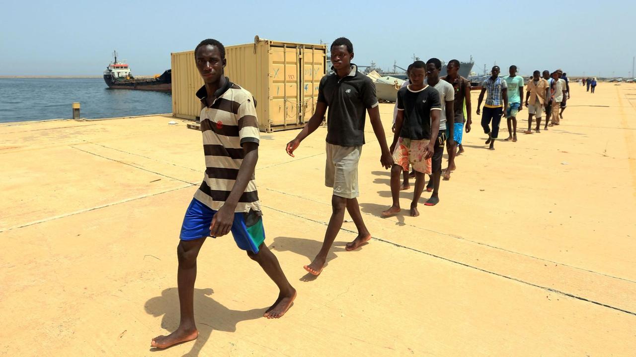 Migranten aus Afrika gehen im Hafen von Tripolis in einer Reihe hintereinander her.