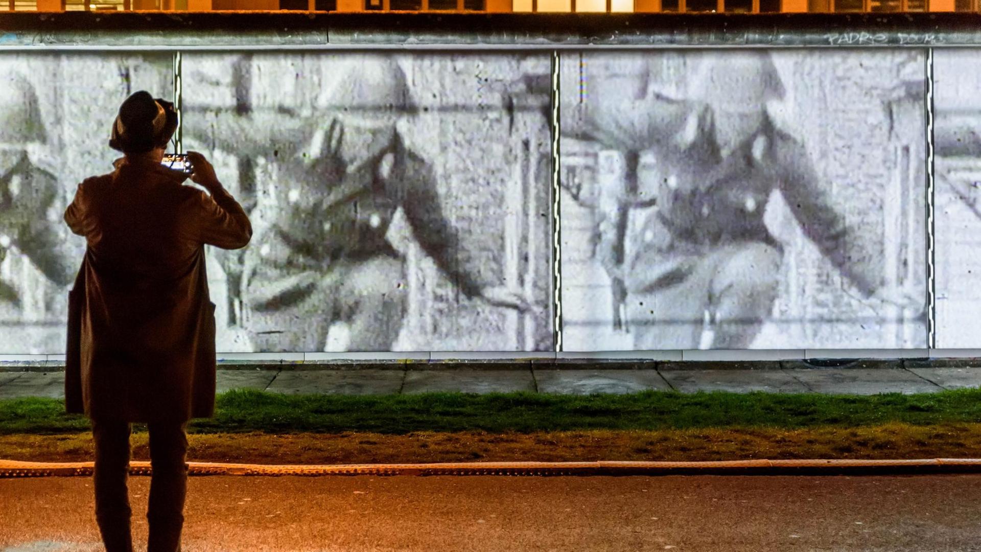 Open-Air-Ausstellung an der East Side Gallery zum 30-jährigen Jubiläum des Falls der Berliner Mauer. Ein Besucher fotografiert eine Projektion der berühmten Grenzflucht des DDR-Soldaten Conrad Schumann, der über einen Stacheldraht springt und nach West-Berlin entkommt.