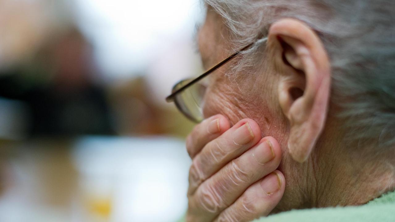 Der Hinterkopf einer alten weißhaarigen Frau, die ihren Kopf auf die linke Hand stützt.