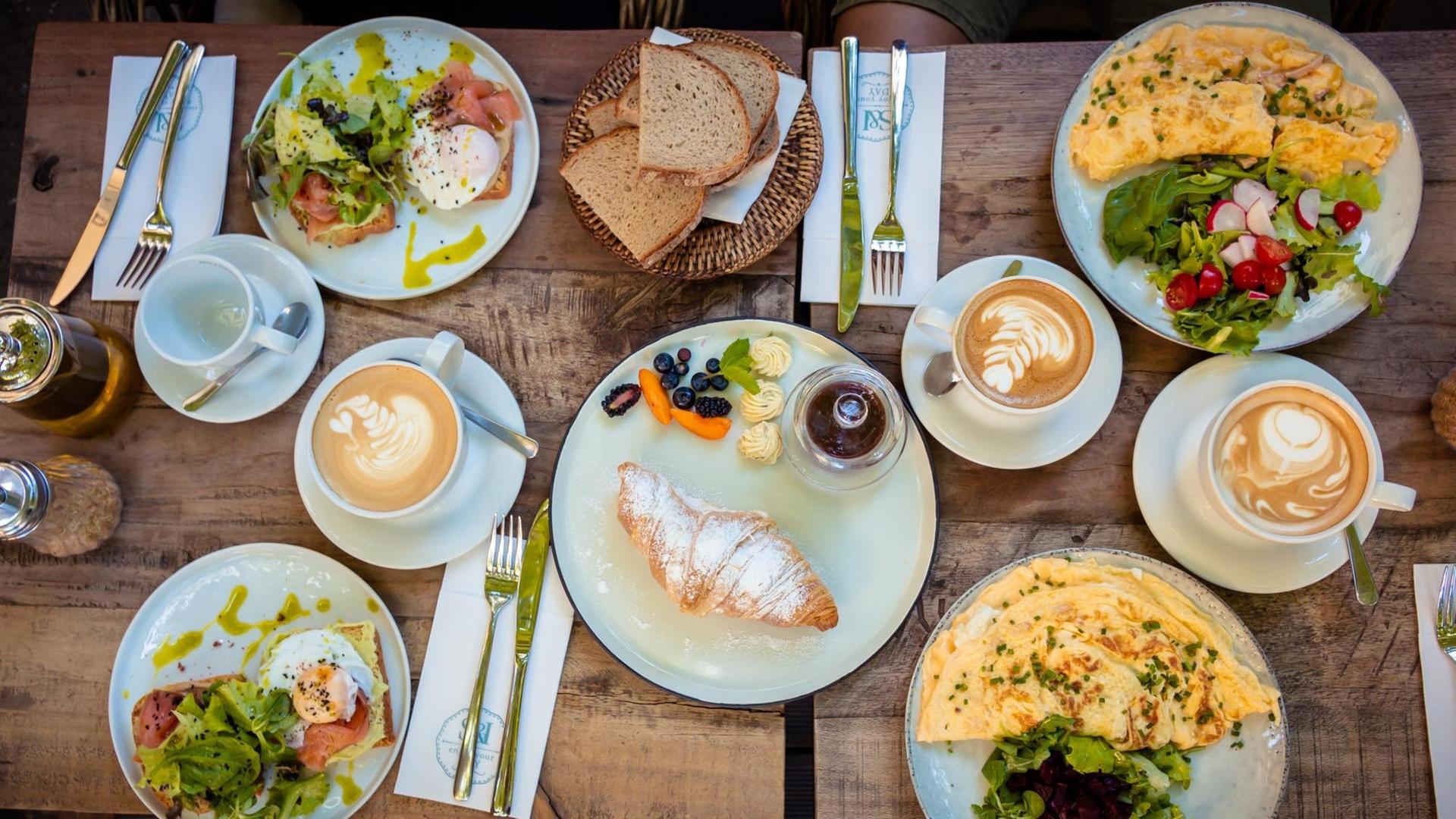 Das Foto zeigt einen gedeckten Frühstückstisch von oben. Auf vier Tellern sind Speisen angerichtet, unter anderem Rührei, Brot und ein Croissant mit Puderzucker sind zu sehen sowie Milchkaffee in Tassen.