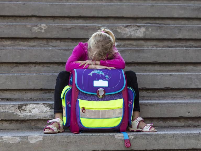 Ein kleines Mädchen sitzt auf Stufen, den Kopf in ihren Schulranzen vergraben.