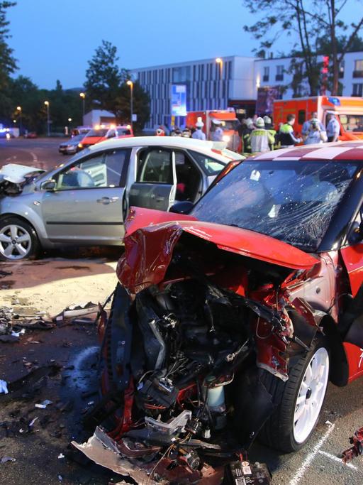 Das Bild zeigt zwei schwer beschädigte Autos an der Unfallstelle. Im Hintergrund zwei Rettungswagen, Sanitäter, Feuerwehrleute und Polizisten.