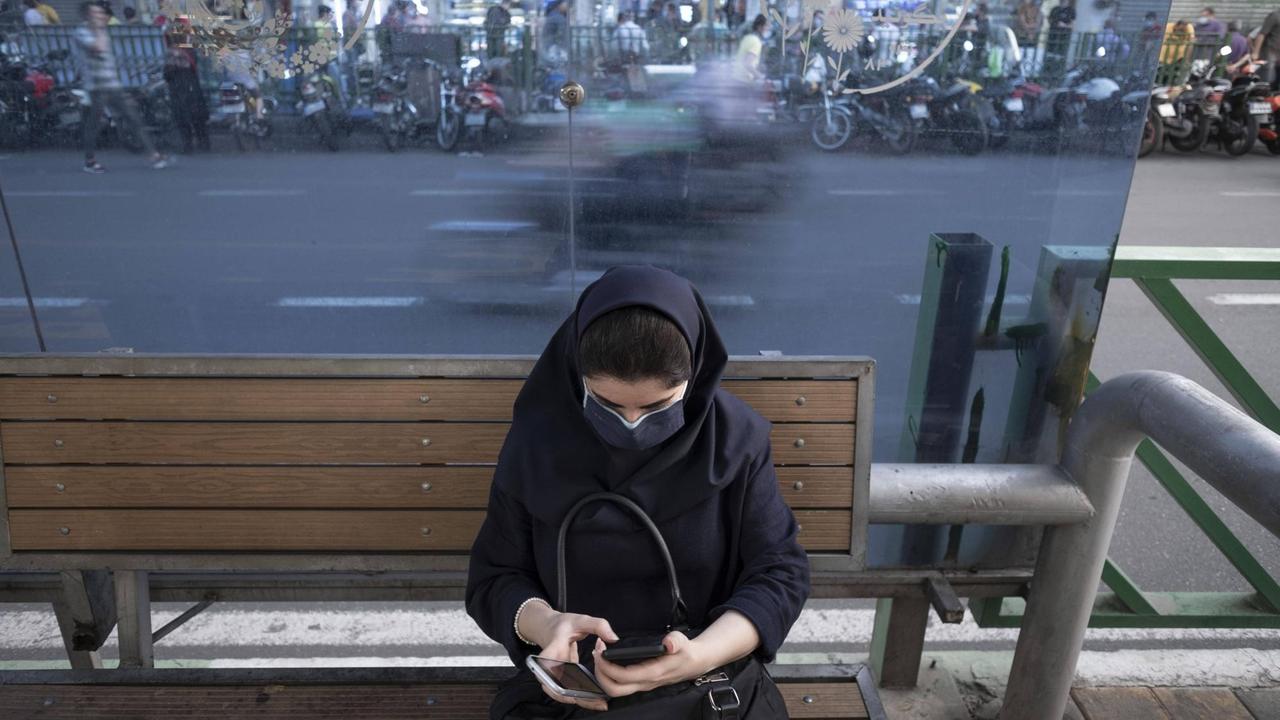 Ein Frau mit dunklem Kopftuch, dunkler Jacke und Mund-Nasen-Schutz sitzt auf einer Bank an einer Straße und schaut auf das Display eines Smartphones.