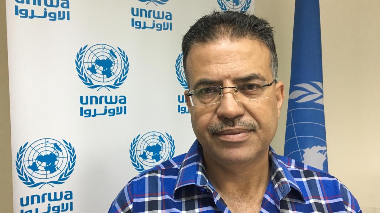 Adnan Abu Hasna, Medienberater/Sprecher des Hilfswerks der Vereinten Nationen für Palästina (UNRWA) steht vor einem Banner mit dem Logo der  der Vereinten Nationen darauf. Im Hintergrund steht außerdem eine Fahne der Organisation.