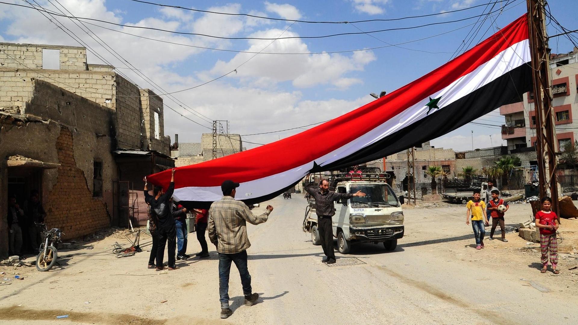 Menschen halten auf einer staubigen Straße eine große syrische Flagge hoch.