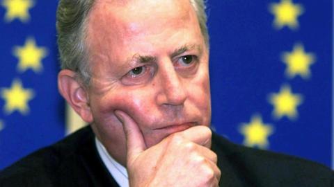 Jacques Santer bei einer Pressekonferenz 1999. Im Hintergrund ist eine EU-Flagge zu sehen.