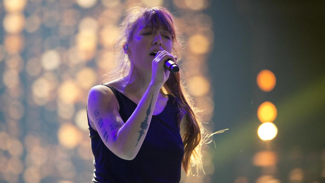 Die Sängerin steht auf der Bühne, ein Mikrofon in der Hand. Im Hintergrund Bühnenbeleuchtung.