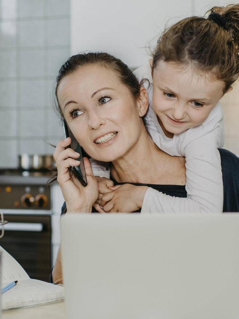 Mutter bei Homeoffice in der Küche, mit Kind am Hals und Haushalt um sich herum. Sie telefoniert und hat einen Laptop vor sich am Küchentisch.(Symbolbild)
