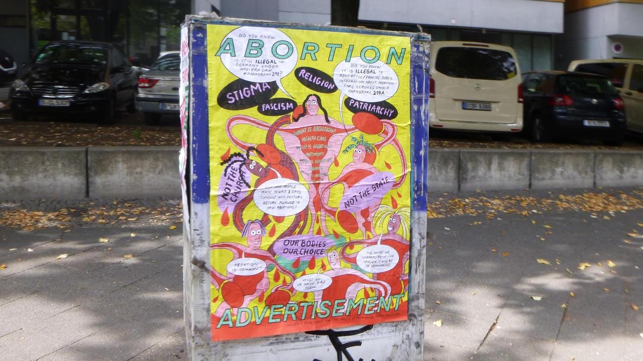 Am Stromkasten auf der Straße klebt ein Plakat. Auf dem illustrierten Motiv sind unter anderem die Wörter "Abortion" und "Adverstisesent" zu sehen.