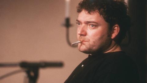 Wiglaf Droste sitzt auf einer Bühne mit Zigarette im Mund. Im Hintergrund steht ein Mikrofon.