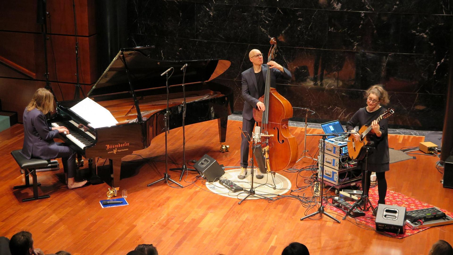 Von links nach rechts sind ein Pianist am Flügel, ein Kontrabassist und eine Gitarristin von schräg oben beim Spielen vor Publikum zu sehen. Sie stehen ebenerdig auf Parkettboden. Der Pianist trägt die langen Haare offen und ist barfuß. Von links nach rechts sind ein Pianist am Flügel, ein Kontrabassist und eine Gitarristin von schräg oben beim Spielen vor Publikum zu sehen. Sie stehen ebenerdig auf Parkettboden. Der Pianist trägt die langen Haare offen und ist barfuß.