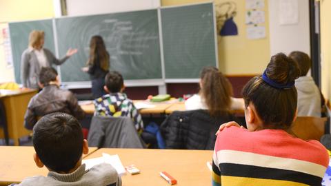 Kinder sitzen in einem Klassenraum und hören der Lehrerin zu, die etwas an der Tafel erklärt.