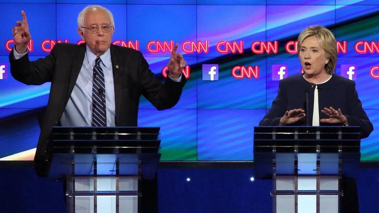 Bernie Sanders und Hillary Clinton reden und gestikulieren an drei Stehpulten im CNN-Studio.