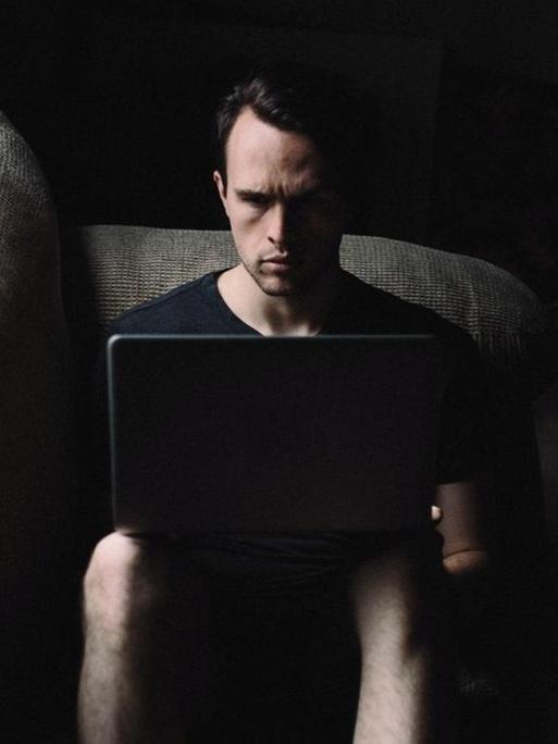 Ein Mann sitzt in einem Zimmer in einem Sessel mit einem Laptop auf den Knien.