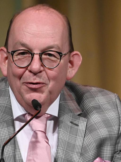 Denis Scheck mit kariertem Jackett und rosa Krawatte lächend an einem Mikrofon.