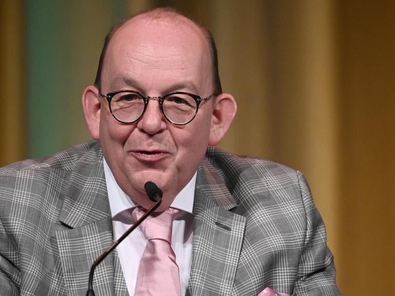 Denis Scheck mit kariertem Jackett und rosa Krawatte lächend an einem Mikrofon.